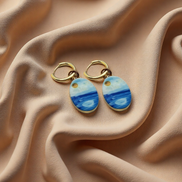 A pair of gold hoop earrings with oval charms painted with a whimsical watercolor style seascape, featuring shades of blue and white with a small gold sun detail, displayed on a beige fabric background. The hoops are PVD 18K Gold Coated Titanium.