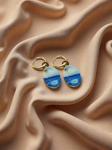 A pair of gold hoop earrings with oval charms painted with a whimsical watercolor style seascape, featuring shades of blue and white with a small gold sun detail, displayed on a beige fabric background. The hoops are PVD 18K Gold Coated Titanium.