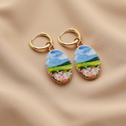 A pair of gold hoop earrings with oval charms featuring a colorful landscape painting of a green field, blue sky, and pink flowers, displayed on a beige fabric background.