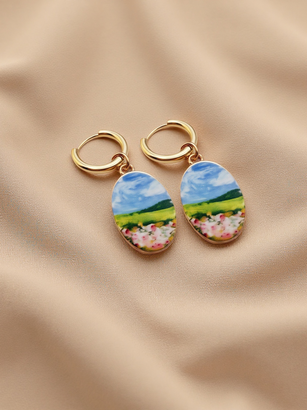 A pair of gold hoop earrings with oval charms featuring a colorful landscape painting of a green field, blue sky, and pink flowers, displayed on a beige fabric background.