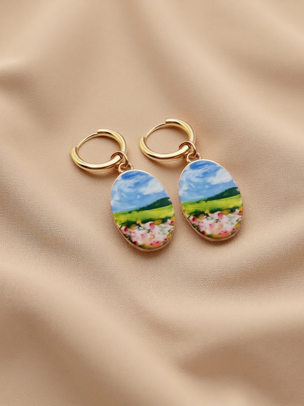 A pair of gold hoop earrings with oval charms featuring a colorful landscape painting of a green field, blue sky, and pink flowers, displayed on a beige fabric background.