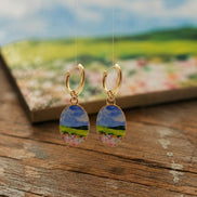 A pair of gold hoop earrings with oval charms depicting a colorful landscape scene of a blue sky, green hills, and pink flowers, displayed on a rustic wooden surface with a blurred scenic background.