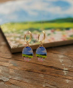 A pair of gold hoop earrings with oval charms depicting a colorful landscape scene of a blue sky, green hills, and pink flowers, displayed on a rustic wooden surface with a blurred scenic background.