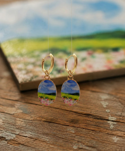 A pair of gold hoop earrings with oval charms depicting a colorful landscape scene of a blue sky, green hills, and pink flowers, displayed on a rustic wooden surface with a blurred scenic background.