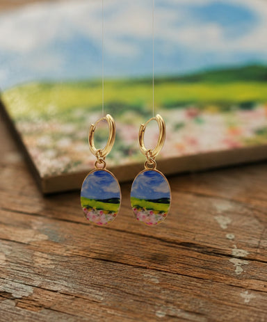 A pair of gold hoop earrings with oval charms depicting a colorful landscape scene of a blue sky, green hills, and pink flowers, displayed on a rustic wooden surface with a blurred scenic background.