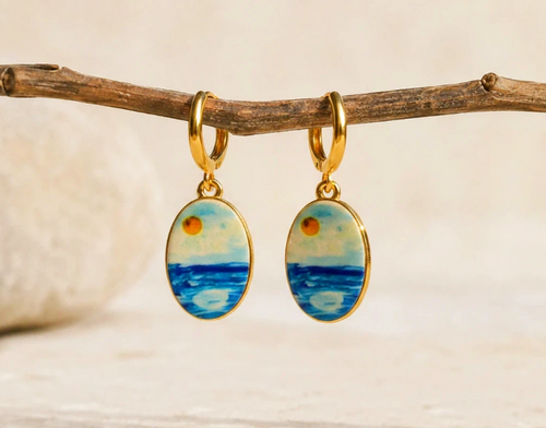 A pair of gold hoop earrings with oval charms featuring a painted seascape scene, hanging from a rustic wooden branch against a soft beige background.