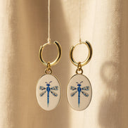 A pair of gold hoop earrings with oval white enamel pendants featuring a delicate and whimsical blue dragonfly design, displayed suspended in front of a beige fabric background. The hoops are PVD 18K Gold Coated Titanium.