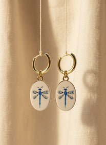 A pair of gold hoop earrings with oval white enamel pendants featuring a delicate and whimsical blue dragonfly design, displayed suspended in front of a beige fabric background. The hoops are PVD 18K Gold Coated Titanium.