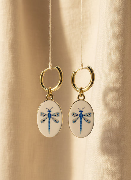 A pair of gold hoop earrings with oval white enamel pendants featuring a delicate and whimsical blue dragonfly design, displayed suspended in front of a beige fabric background. The hoops are PVD 18K Gold Coated Titanium.