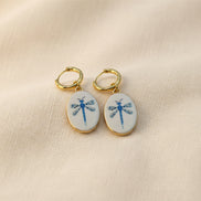 A pair of gold hoop earrings with oval white enamel pendants featuring a delicate and whimsical blue dragonfly design, displayed on a beige fabric background. The hoops are PVD 18K Gold Coated Titanium.