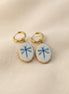A pair of gold hoop earrings with oval white enamel pendants featuring a delicate and whimsical blue dragonfly design, displayed on a beige fabric background. The hoops are PVD 18K Gold Coated Titanium.