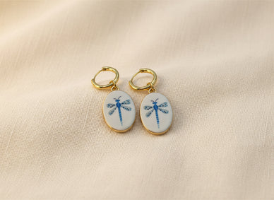 A pair of gold hoop earrings with oval white enamel pendants featuring a delicate and whimsical blue dragonfly design, displayed on a beige fabric background. The hoops are PVD 18K Gold Coated Titanium.