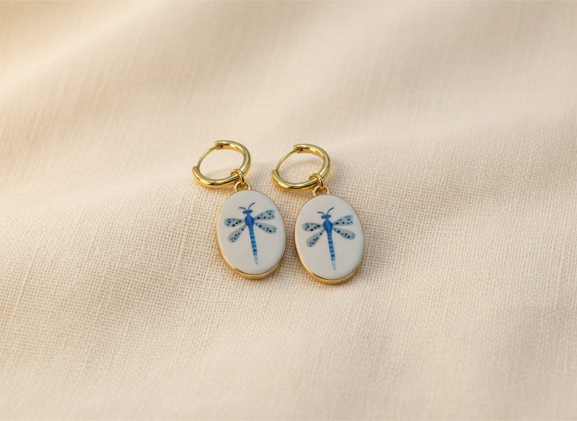 A pair of gold hoop earrings with oval white enamel pendants featuring a delicate and whimsical blue dragonfly design, displayed on a beige fabric background. The hoops are PVD 18K Gold Coated Titanium.