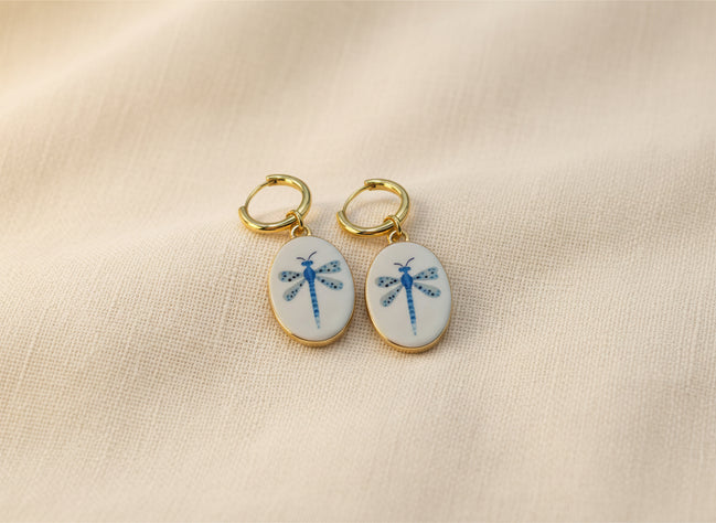 A pair of gold hoop earrings with oval white enamel pendants featuring a delicate and whimsical blue dragonfly design, displayed on a beige fabric background. The hoops are PVD 18K Gold Coated Titanium.
