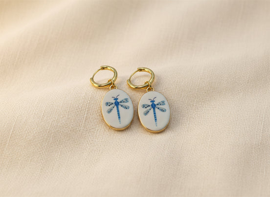 A pair of gold hoop earrings with oval white enamel pendants featuring a delicate and whimsical blue dragonfly design, displayed on a beige fabric background. The hoops are PVD 18K Gold Coated Titanium.