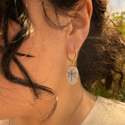 A close-up side-profile portrait showing a person’s ear, cheek, and dark wavy hair in warm golden-hour sunlight. They wear a small handmade gold hoop earring with a dangling oval dragonfly pendant and a delicate gold necklace, with greenery softly blurred in the background. The hoops are 18k gold coated titanium