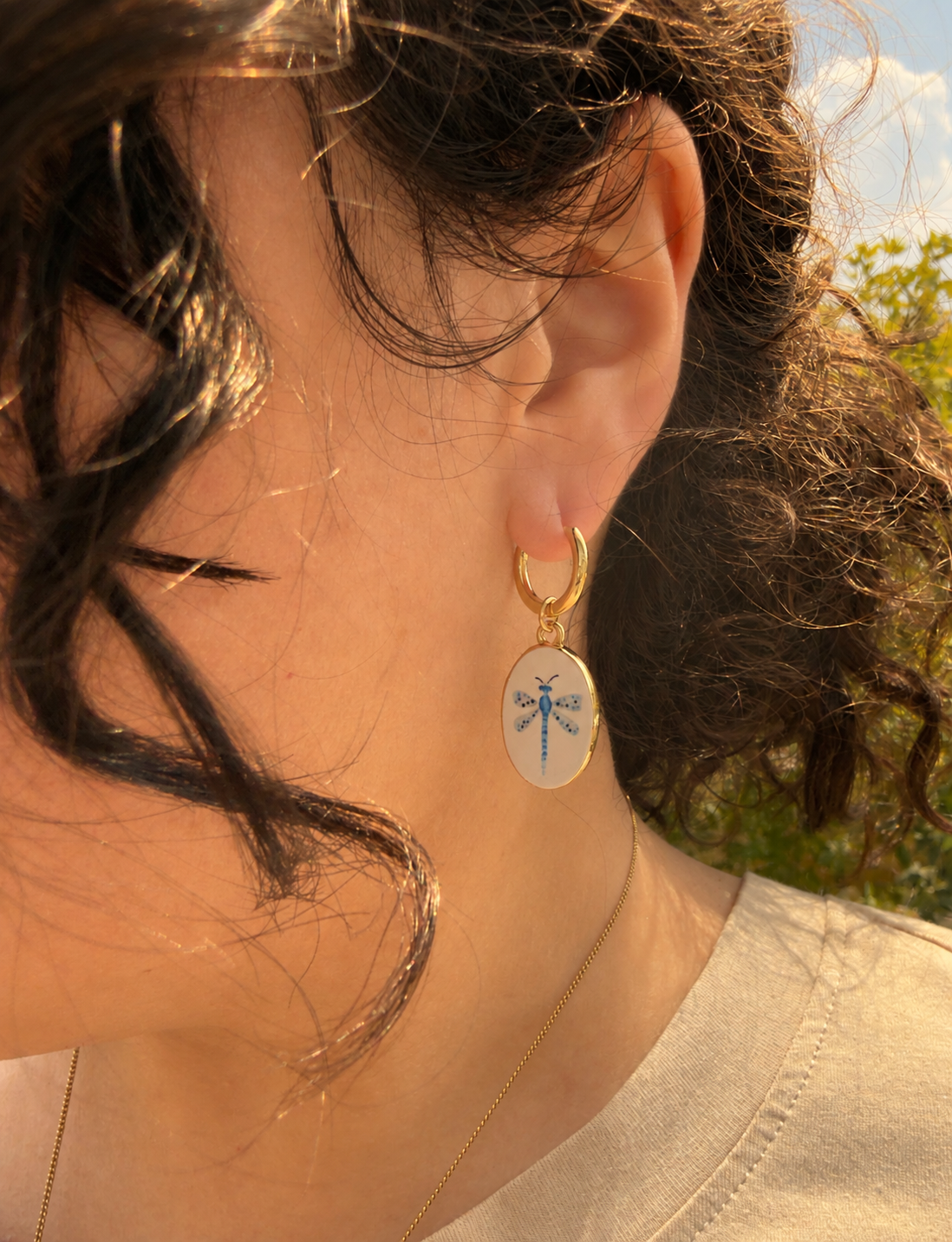 A close-up side-profile portrait showing a person’s ear, cheek, and dark wavy hair in warm golden-hour sunlight. They wear a small handmade gold hoop earring with a dangling oval dragonfly pendant and a delicate gold necklace, with greenery softly blurred in the background. The hoops are 18k gold coated titanium