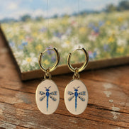 A pair of gold hoop earrings with oval white enamel pendants featuring a delicate and whimsical blue dragonfly design, displayed hanging above a rustic wooden table with an out of focus watercolor painting in the background featuring a field of white and blue flowers below a blue sky. The hoops are PVD 18K Gold Coated Titanium.