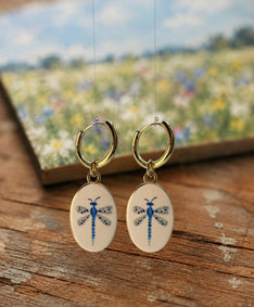 A pair of gold hoop earrings with oval white enamel pendants featuring a delicate and whimsical blue dragonfly design, displayed hanging above a rustic wooden table with an out of focus watercolor painting in the background featuring a field of white and blue flowers below a blue sky. The hoops are PVD 18K Gold Coated Titanium.
