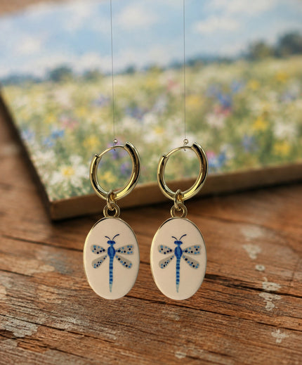 A pair of gold hoop earrings with oval white enamel pendants featuring a delicate and whimsical blue dragonfly design, displayed hanging above a rustic wooden table with an out of focus watercolor painting in the background featuring a field of white and blue flowers below a blue sky. The hoops are PVD 18K Gold Coated Titanium.