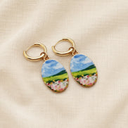 A pair of gold hoop earrings with oval charms featuring a whimsical, watercolor landscape print of green hills, blue sky, and pink flowers, displayed on a textured beige fabric background. The hoops are PVD 18K Gold Coated titanium.