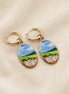 A pair of gold hoop earrings with oval charms featuring a whimsical, watercolor landscape print of green hills, blue sky, and pink flowers, displayed on a textured beige fabric background. The hoops are PVD 18K Gold Coated titanium.