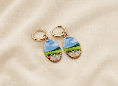 A pair of gold hoop earrings with oval charms featuring a whimsical, watercolor landscape print of green hills, blue sky, and pink flowers, displayed on a textured beige fabric background. The hoops are PVD 18K Gold Coated titanium.
