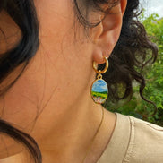 A close-up of a person wearing titanium hoops and brass charms finished with PVD 18K gold plating. The charm features a whimsical watercolor-inspired landscape: a field of soft pink flowers stretching toward a peaceful meadow, with a distant mountain beneath an open blue sky.