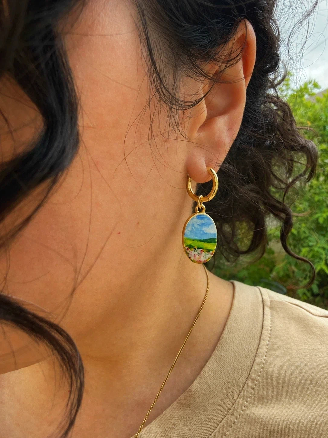 A close-up of a person wearing titanium hoops and brass charms finished with PVD 18K gold plating. The charm features a whimsical watercolor-inspired landscape: a field of soft pink flowers stretching toward a peaceful meadow, with a distant mountain beneath an open blue sky.