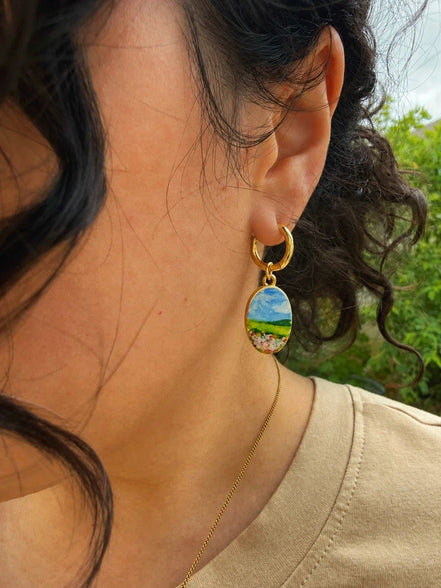 A close-up of a person wearing titanium hoops and brass charms finished with PVD 18K gold plating. The charm features a whimsical watercolor-inspired landscape: a field of soft pink flowers stretching toward a peaceful meadow, with a distant mountain beneath an open blue sky.