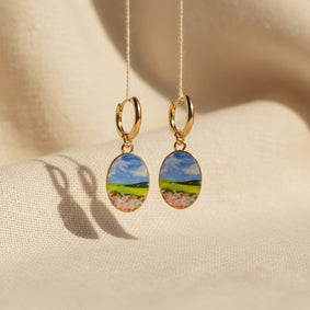 A pair of gold hoop earrings with oval charms featuring a whimsical, watercolor landscape print of green hills, blue sky, and pink flowers, suspended in front of a textured beige fabric background. The hoops are PVD 18K Gold Coated titanium.