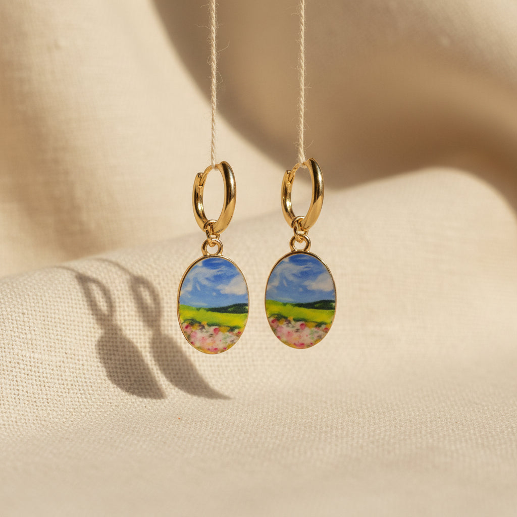 A pair of gold hoop earrings with oval charms featuring a whimsical, watercolor landscape print of green hills, blue sky, and pink flowers, suspended in front of a textured beige fabric background. The hoops are PVD 18K Gold Coated titanium.
