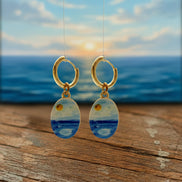 A pair of gold hoop earrings with oval dangles featuring a blue and yellow seascape design, shown against a blurred ocean sunset background with a wooden surface in the foreground.