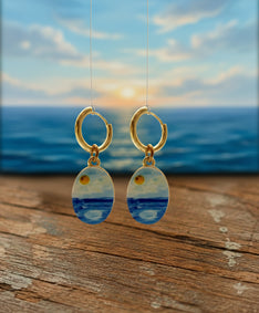 A pair of gold hoop earrings with oval dangles featuring a blue and yellow seascape design, shown against a blurred ocean sunset background with a wooden surface in the foreground.