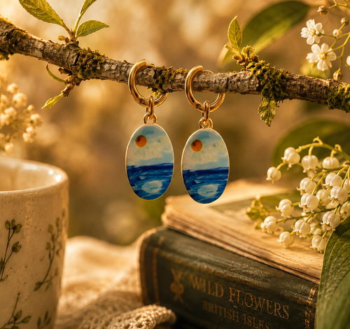 A pair of oval drop earrings with gold-tone rings and blue-and-white seascape charms, hanging from a mossy branch in a styled setting with flowers, a mug, and an old book.
