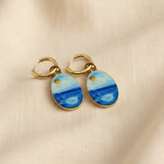 A pair of gold hoop earrings with oval charms painted to look like a seascape, featuring shades of blue and white with a small gold sun detail, displayed on a beige fabric background.