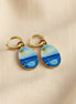 A pair of gold hoop earrings with oval charms painted to look like a seascape, featuring shades of blue and white with a small gold sun detail, displayed on a beige fabric background.