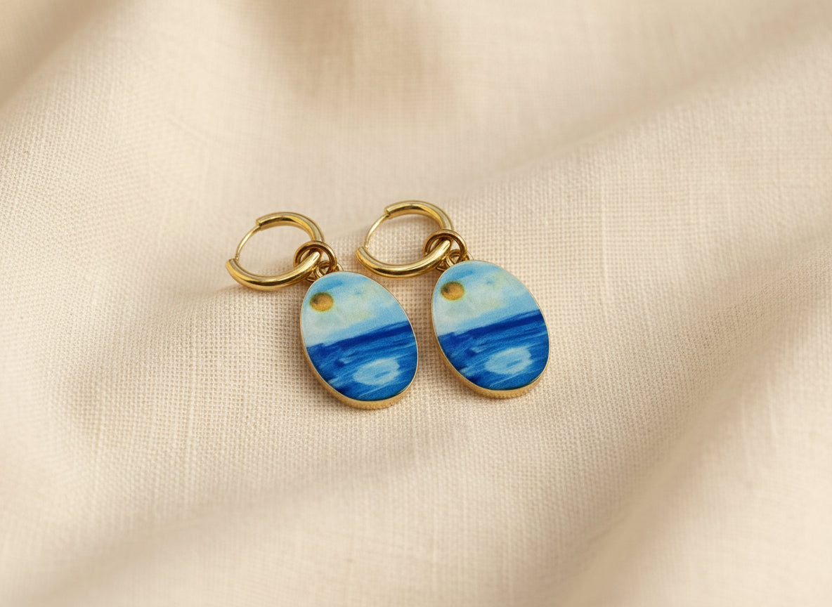 A pair of gold hoop earrings with oval charms painted to look like a seascape, featuring shades of blue and white with a small gold sun detail, displayed on a beige fabric background.