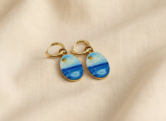 A pair of gold hoop earrings with oval charms painted to look like a seascape, featuring shades of blue and white with a small gold sun detail, displayed on a beige fabric background.