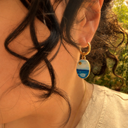 A close-up lifestyle shot of a person wearing a gold huggie hoop earring with a small oval charm depicting a whimsical watercolor inspired ocean and white clouds landscape with a yellow sun, paired with a gold chain necklace. The hoops are PVD 18K Gold Coated Titanium.