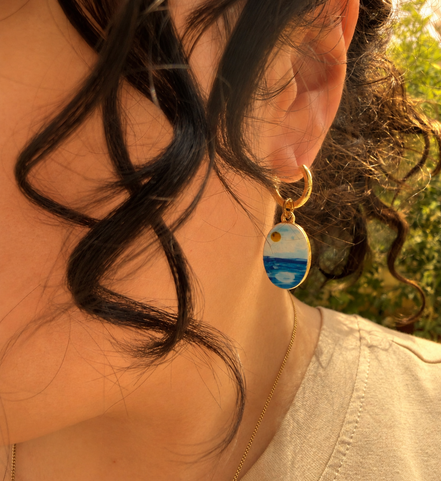 A close-up lifestyle shot of a person wearing a gold huggie hoop earring with a small oval charm depicting a whimsical watercolor inspired ocean and white clouds landscape with a yellow sun, paired with a gold chain necklace. The hoops are PVD 18K Gold Coated Titanium.