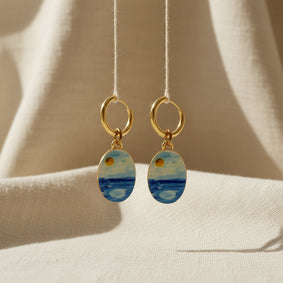 A pair of gold hoop earrings with oval charms painted with a whimsical watercolor style seascape, featuring shades of blue and white with a small gold sun detail, displayed on a beige fabric background. The hoops are PVD 18K Gold Coated Titanium.