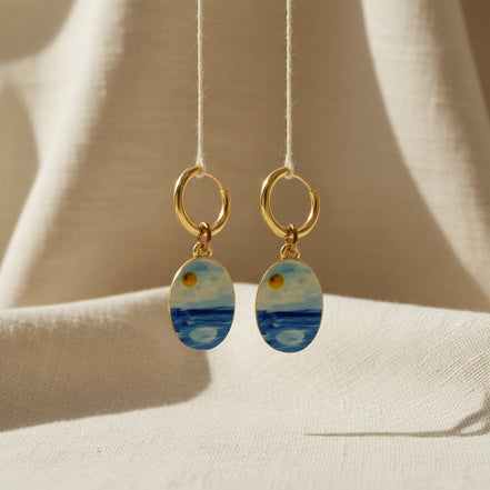 A pair of gold hoop earrings with oval charms painted with a whimsical watercolor style seascape, featuring shades of blue and white with a small gold sun detail, displayed on a beige fabric background. The hoops are PVD 18K Gold Coated Titanium.