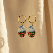 A pair of handmade gold hoop earrings with oval charms featuring a hand-painted watercolor style landscape of red flowers under a blue sky, displayed suspended in front of a textured beige fabric background. The hoops are PVD 18k Gold Coated Titanium.