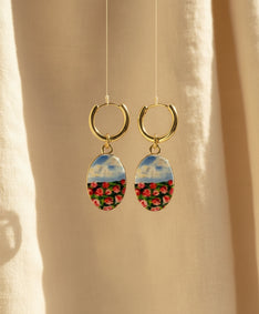 A pair of handmade gold hoop earrings with oval charms featuring a hand-painted watercolor style landscape of red flowers under a blue sky, displayed suspended in front of a textured beige fabric background. The hoops are PVD 18k Gold Coated Titanium.