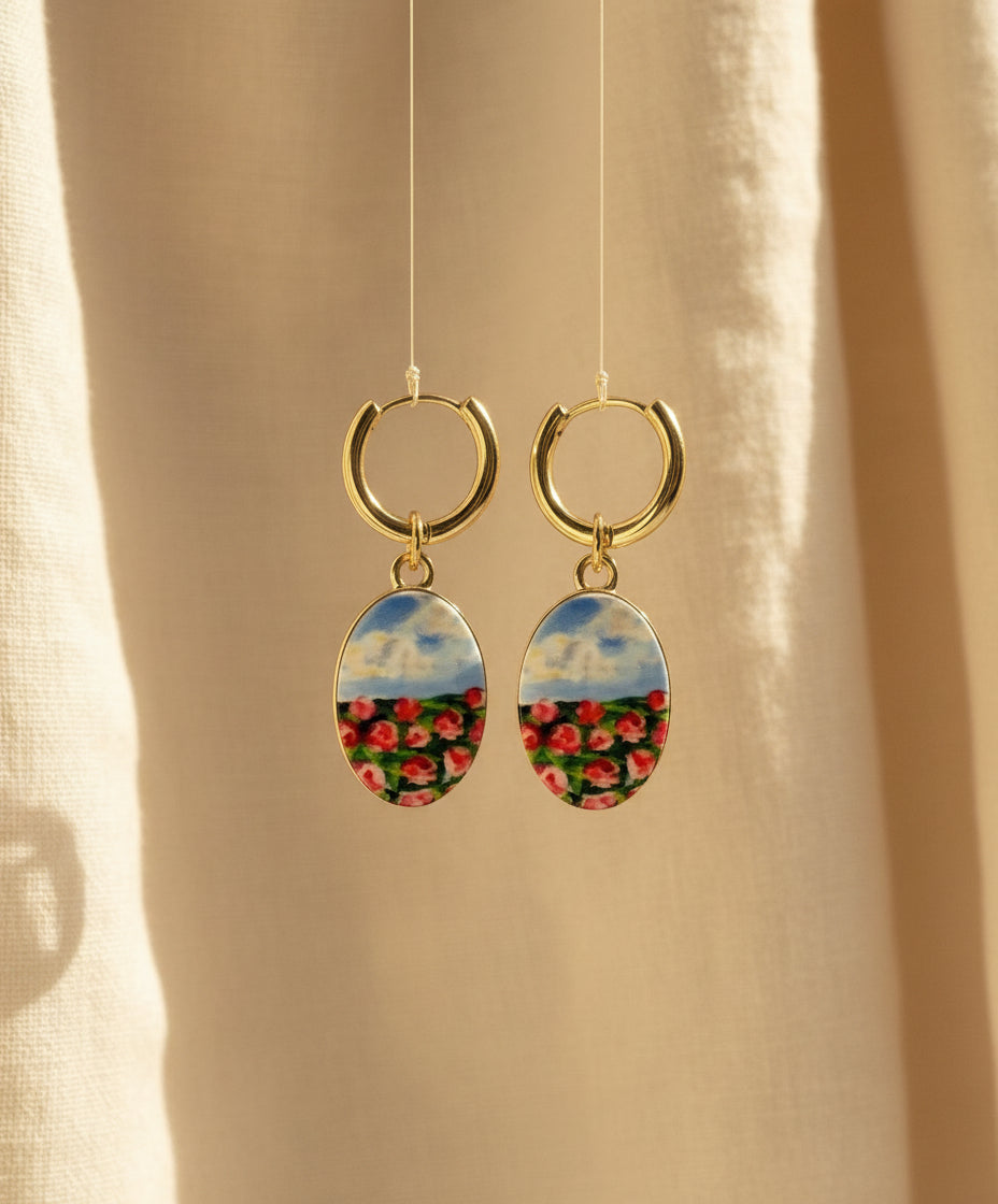 A pair of handmade gold hoop earrings with oval charms featuring a hand-painted watercolor style landscape of red flowers under a blue sky, displayed suspended in front of a textured beige fabric background. The hoops are PVD 18k Gold Coated Titanium.