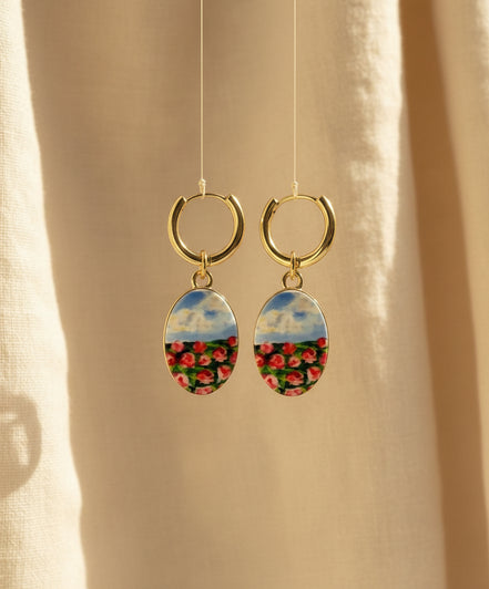 A pair of handmade gold hoop earrings with oval charms featuring a hand-painted watercolor style landscape of red flowers under a blue sky, displayed suspended in front of a textured beige fabric background. The hoops are PVD 18k Gold Coated Titanium.