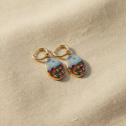 A pair of handmade gold hoop earrings with oval charms featuring a hand-painted watercolor style landscape of red flowers under a blue sky, displayed on a textured beige fabric background. The hoops are PVD 18k Gold Coated Titanium.