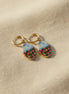 A pair of handmade gold hoop earrings with oval charms featuring a hand-painted watercolor style landscape of red flowers under a blue sky, displayed on a textured beige fabric background. The hoops are PVD 18k Gold Coated Titanium.