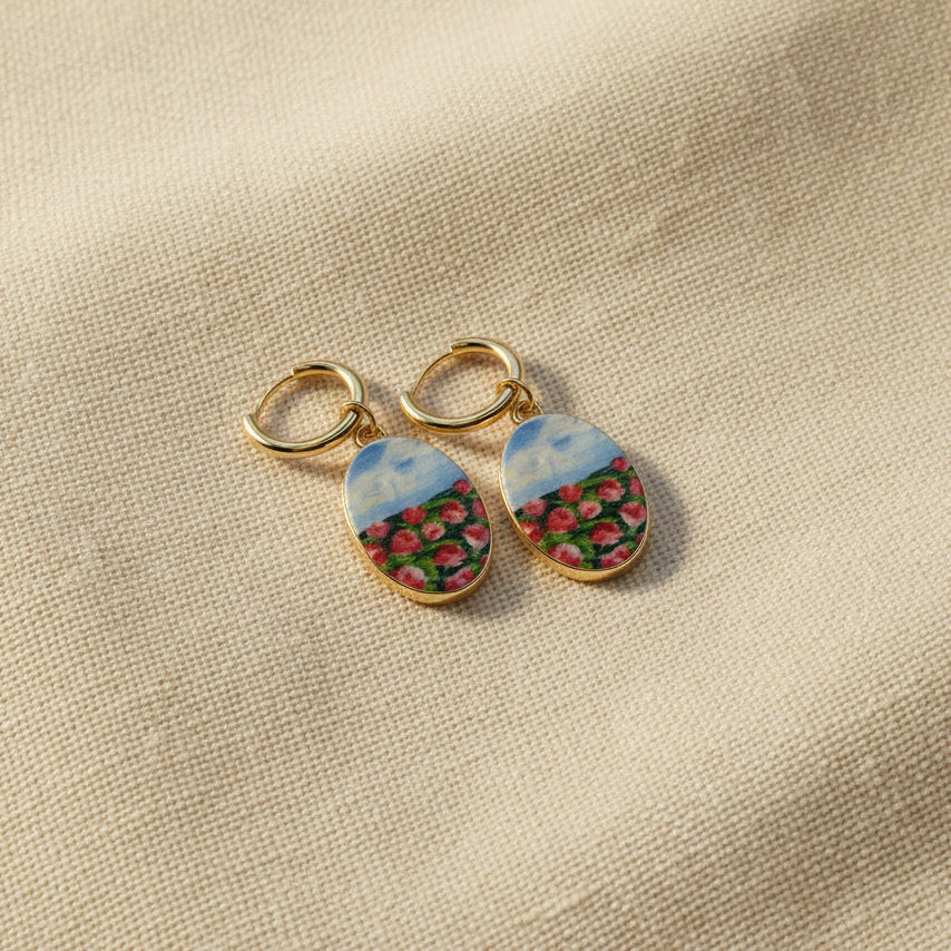 A pair of handmade gold hoop earrings with oval charms featuring a hand-painted watercolor style landscape of red flowers under a blue sky, displayed on a textured beige fabric background. The hoops are PVD 18k Gold Coated Titanium.